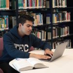 Estudante em uma biblioteca universitária, usando laptop e livros para estudar e fazer pesquisas.