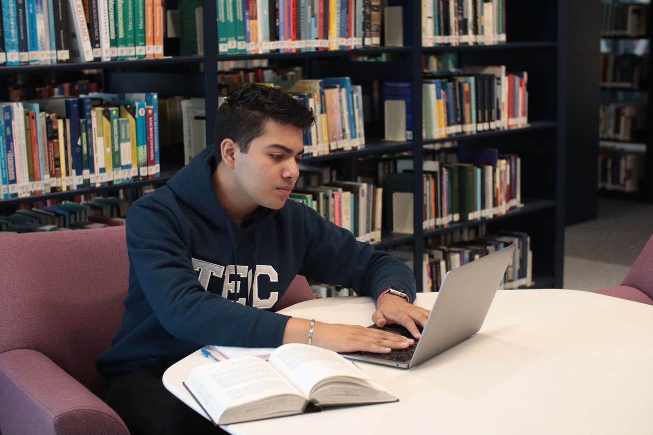 Estudante em uma biblioteca universitária, usando laptop e livros para estudar e fazer pesquisas.