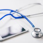 Smartphone ao lado de um estetoscópio azul sobre uma superfície branca, em referência à saúde e ao uso de serviços digitais.