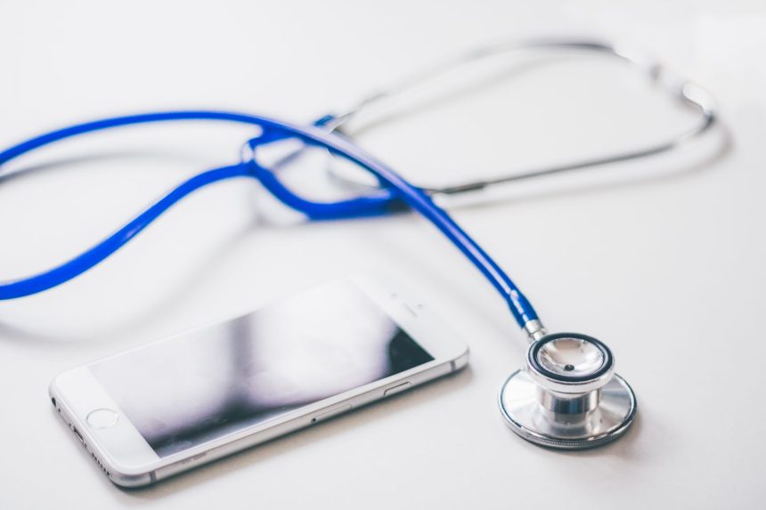 Smartphone ao lado de um estetoscópio azul sobre uma superfície branca, em referência à saúde e ao uso de serviços digitais.